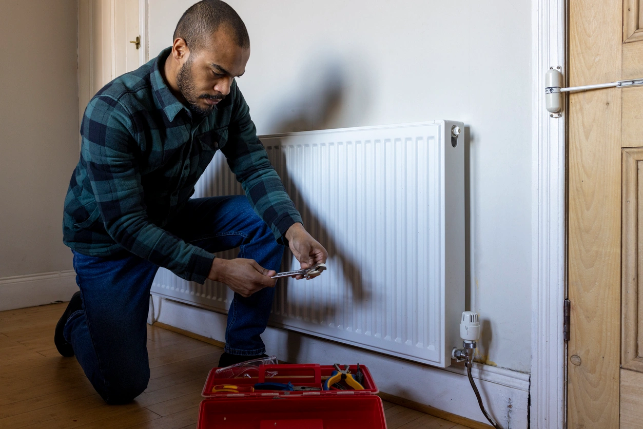 A man repairs a radiator in a room, focusing on the task with tools in hand and a determined expression.